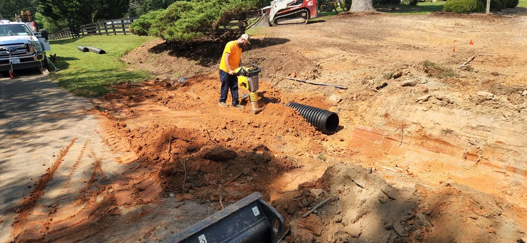 Construction worker using a compactor on a dirt site with equipment and drainage pipe.