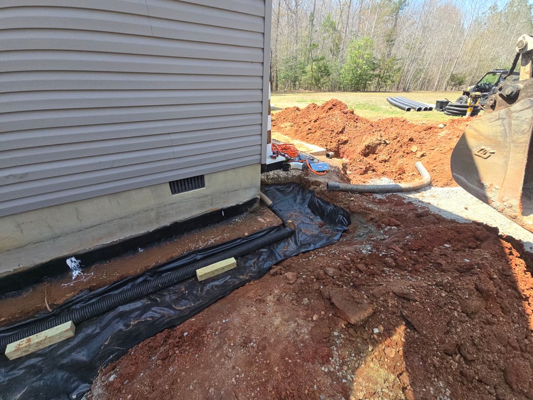 Drainage system installation around home's foundation with excavated earth and construction equipment.