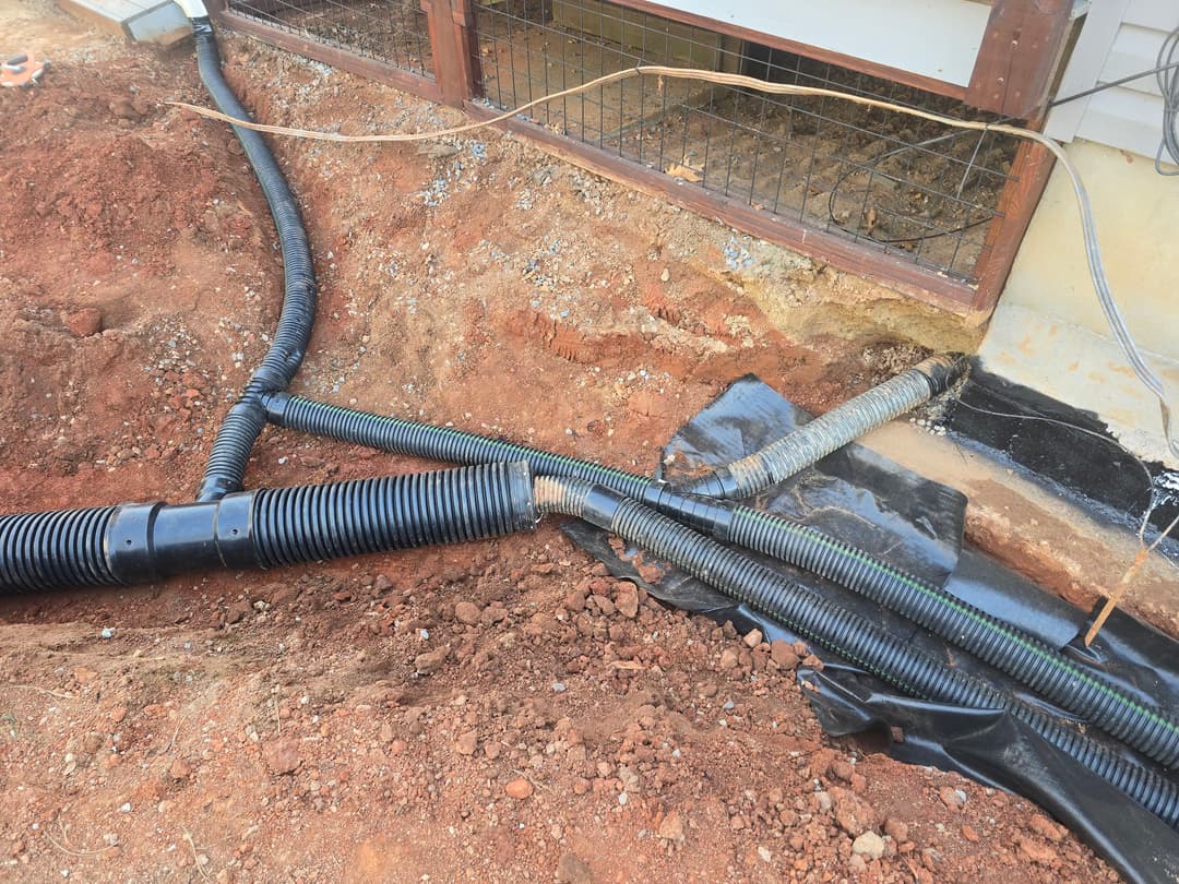 Drainage pipes and black tarp installation near a home's foundation on exposed soil.