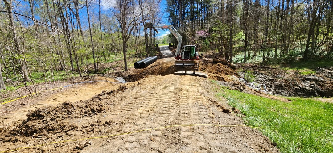 Construction site with machinery, gravel path, and pipe installation in a wooded area.