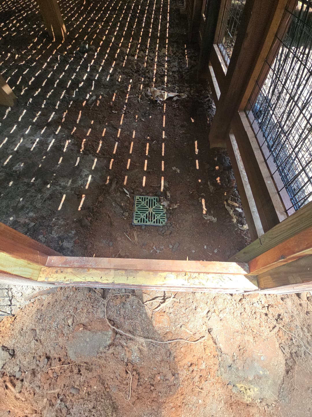 Drainage grate in wooden structure with sunlight casting striped shadows on the ground.