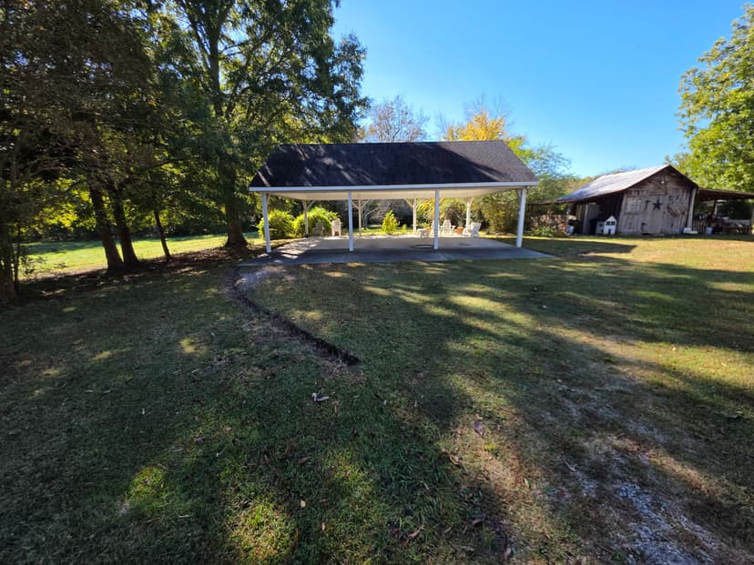 Spacious outdoor pavilion surrounded by trees and a rustic barn in a scenic setting.
