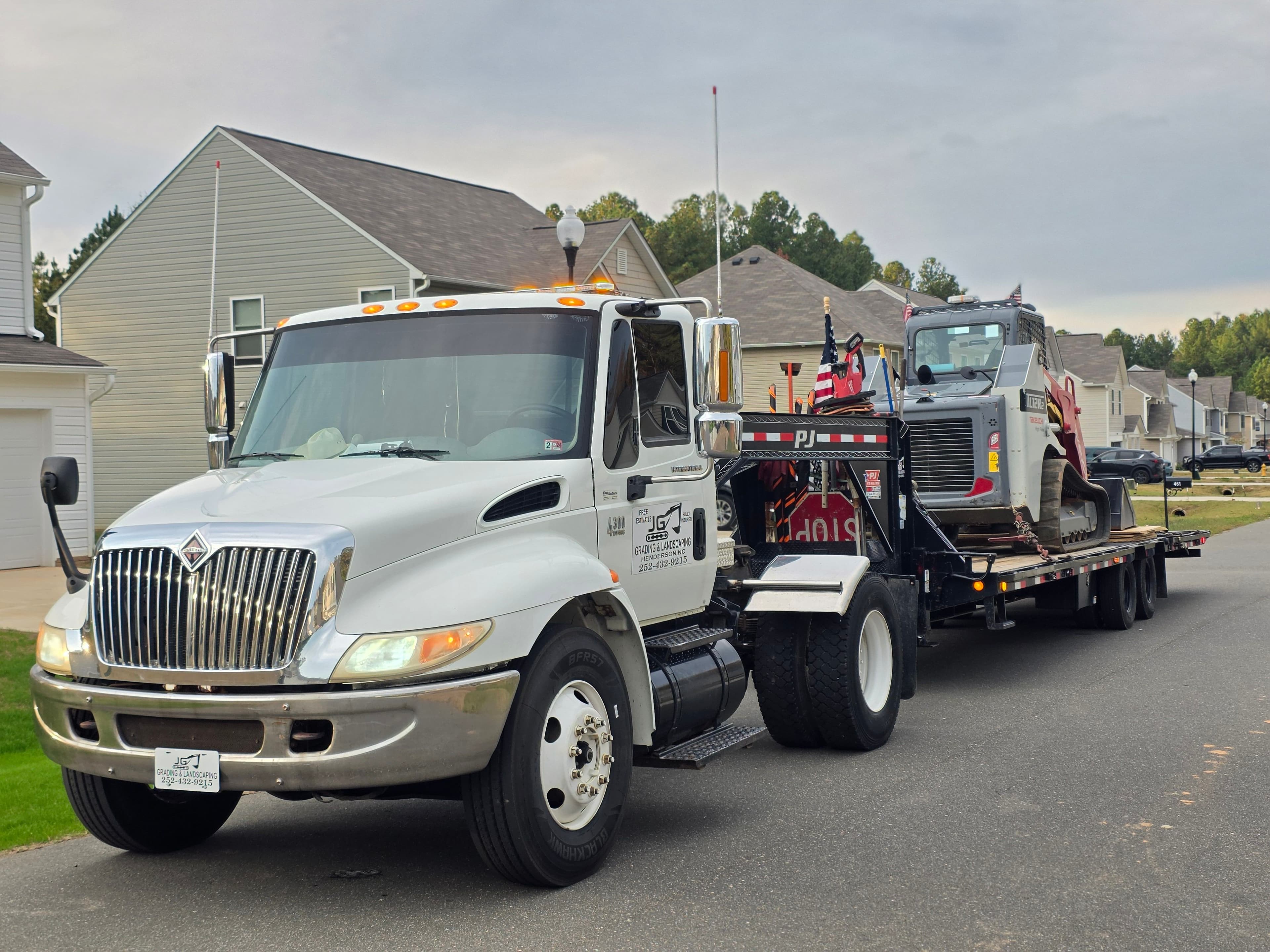 White truck towing construction equipment down a residential street with houses in background.