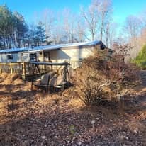 Dilapidated house with wooden deck surrounded by overgrown vegetation and trees.