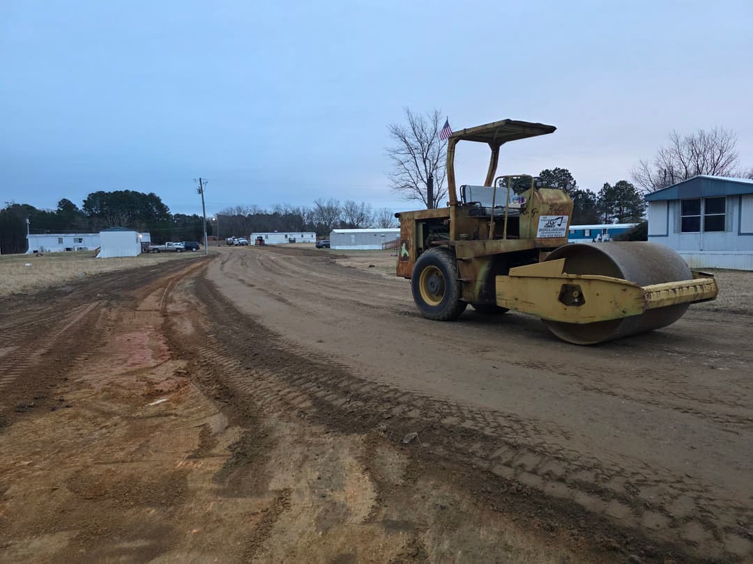 Yellow road roller on dirt road with mobile homes in background during overcast day.