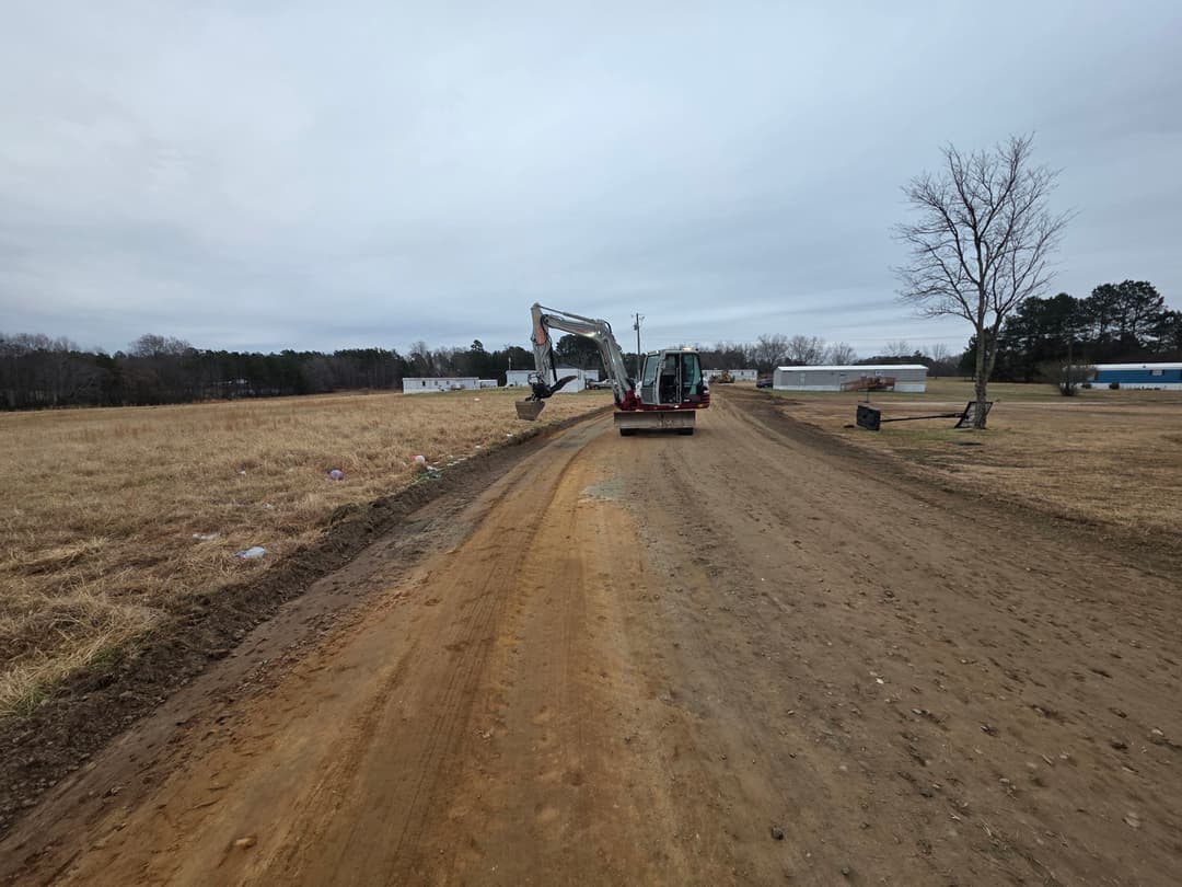 Construction vehicle on a dirt road in a rural area, surrounded by grass and trees.