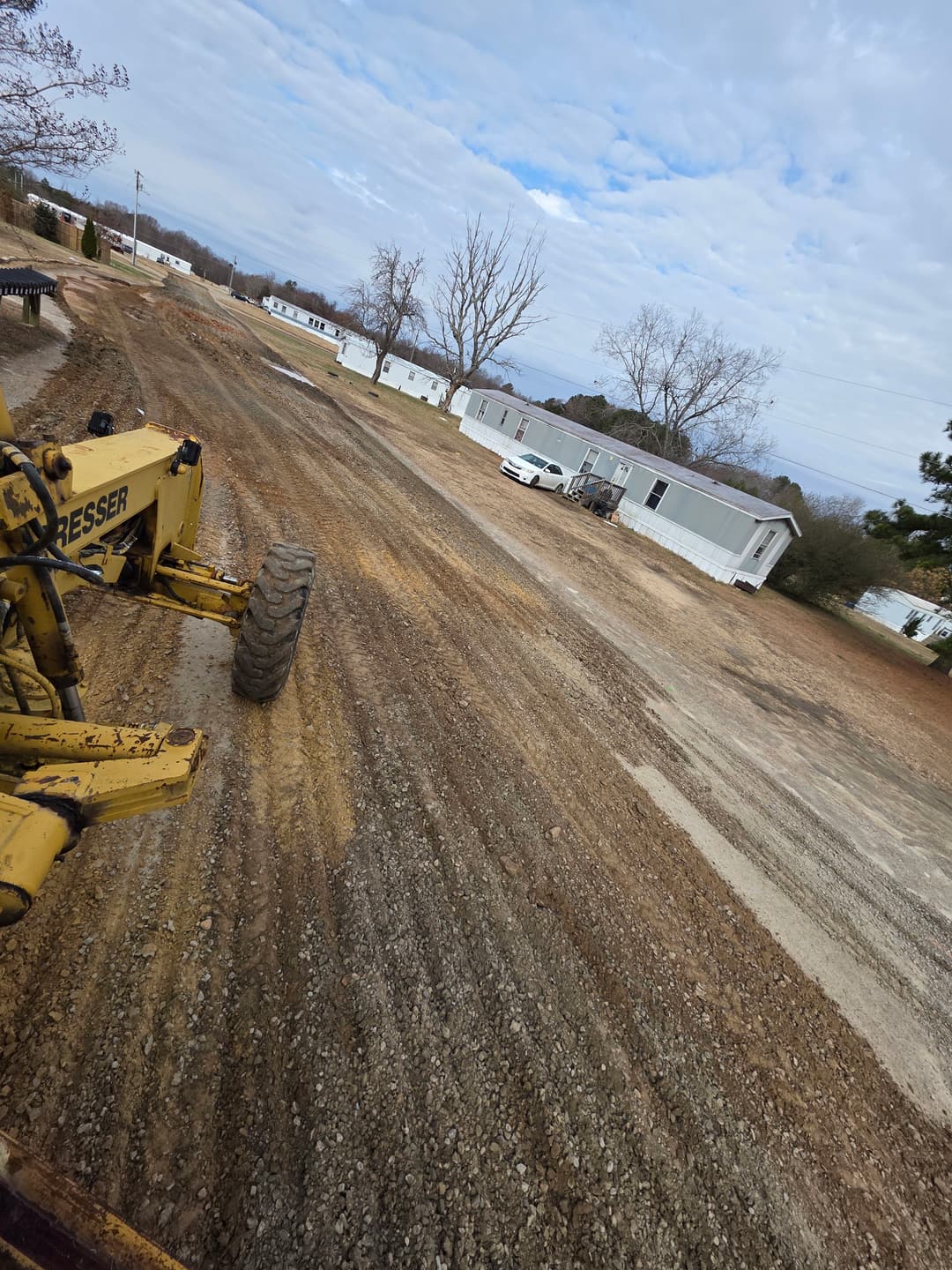 Grader leveling dirt in a mobile home park with cloudy skies and trees in the background.