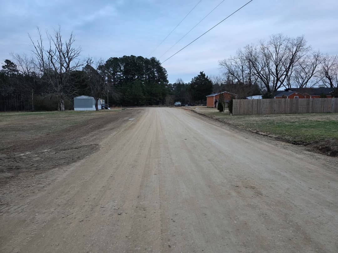 Dirt road lined with trees and buildings under a cloudy sky. Quiet rural scene.