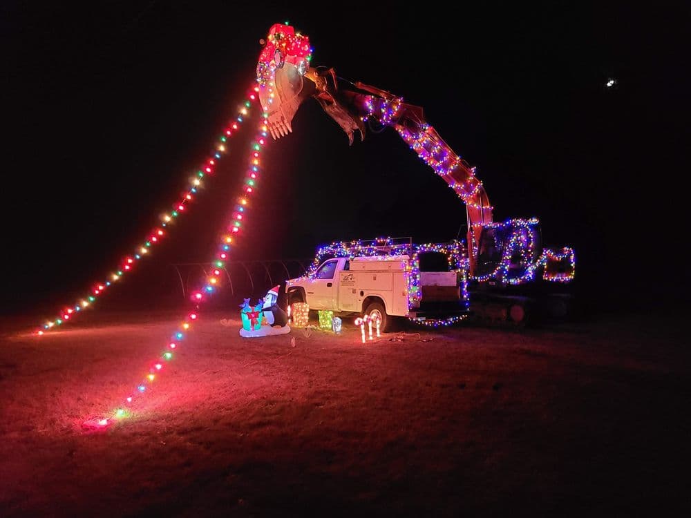 Excavator decorated with colorful Christmas lights in a nighttime display with festive figures.