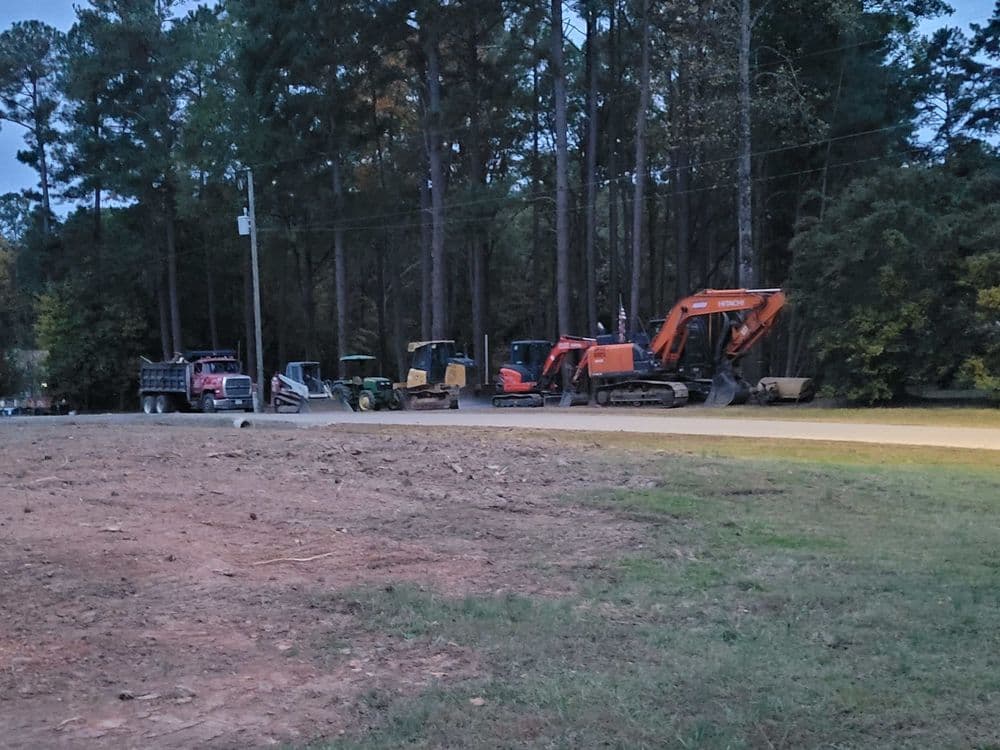 Construction vehicles lined up on a dirt road surrounded by tall trees at dusk.