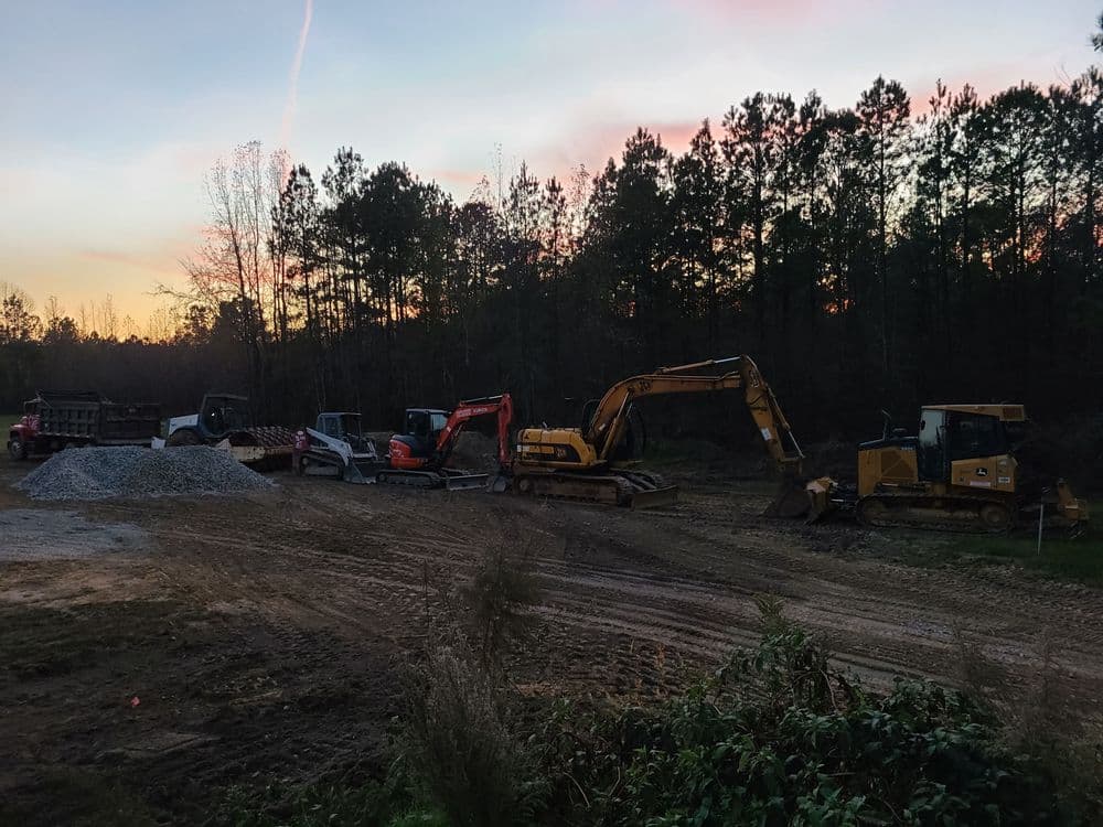 Construction equipment lineup at sunset in a forested area, showcasing heavy machinery.