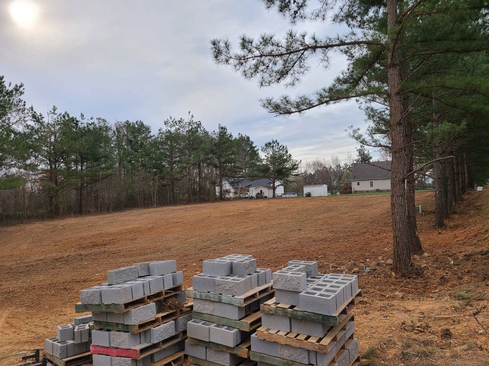 Cleared land with stacked concrete blocks and a forested area near residential buildings.