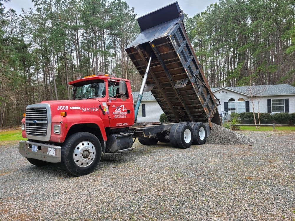 Red dump truck unloading gravel in a residential driveway surrounded by trees.