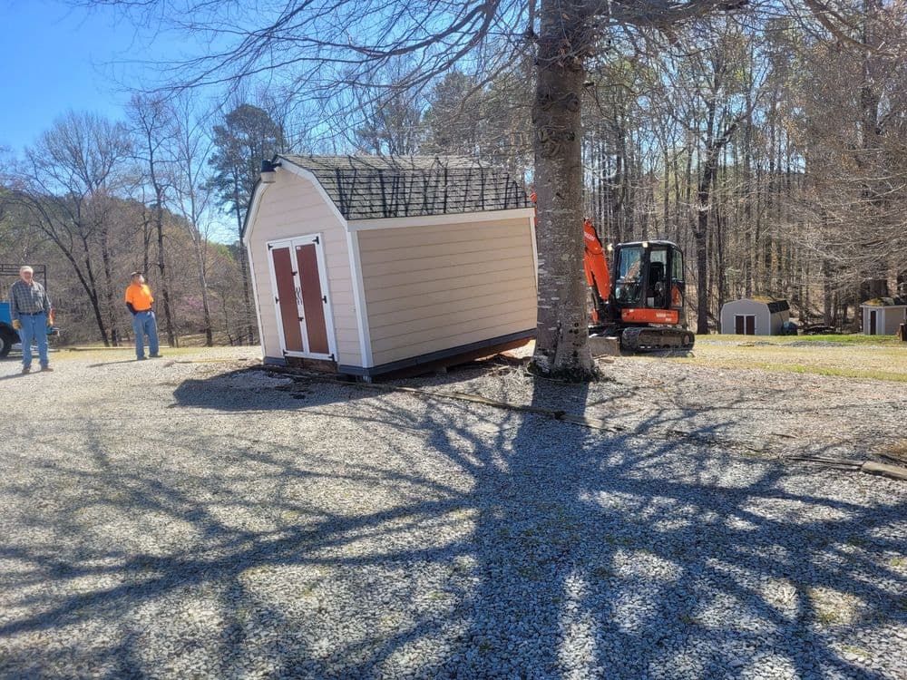 Construction equipment moving a shed in a wooded area, with workers observing nearby.
