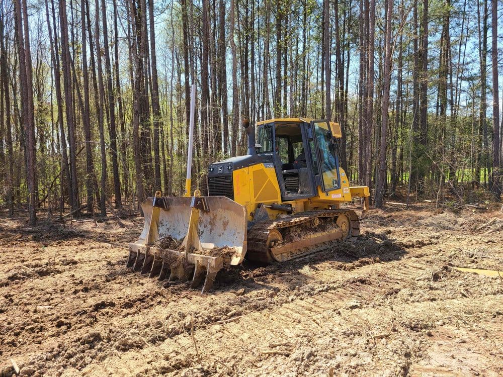 Yellow bulldozer clearing land in a forested area with tall trees.