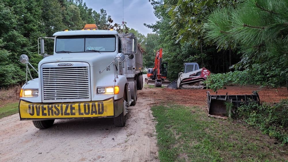Oversized load truck on unpaved road near construction equipment and wooded area.