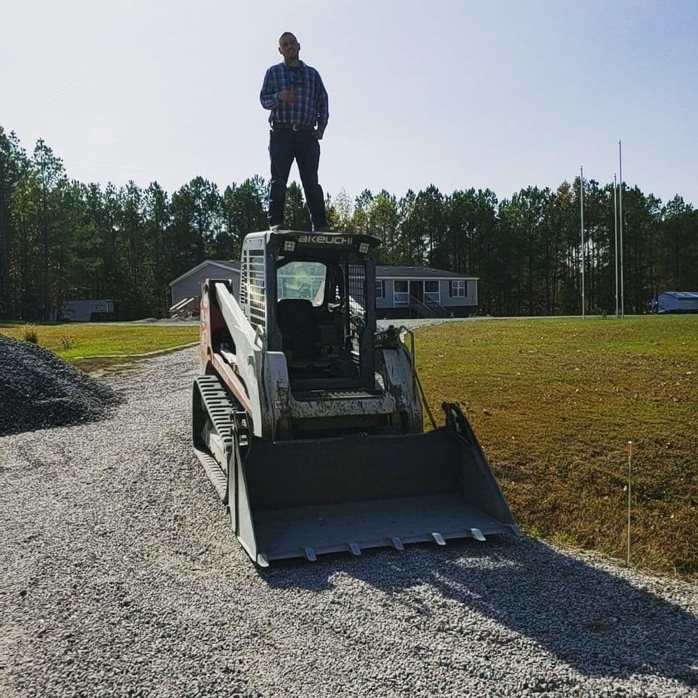 Person standing on a bobcat loader on a gravel path, with trees and a house in the background.