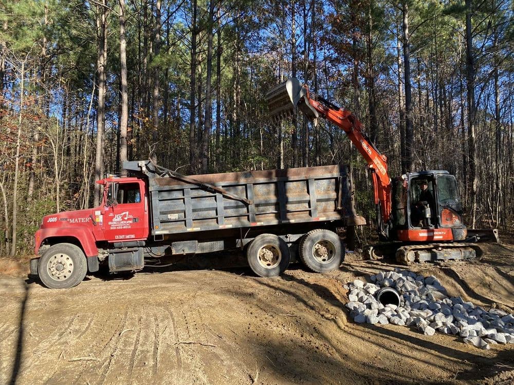 Red dump truck and excavator on construction site surrounded by trees and rocks.