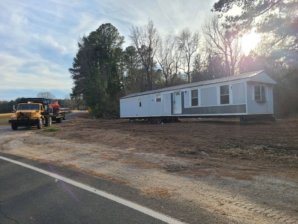 Mobile home being transported on a truck along a rural road with trees in the background.