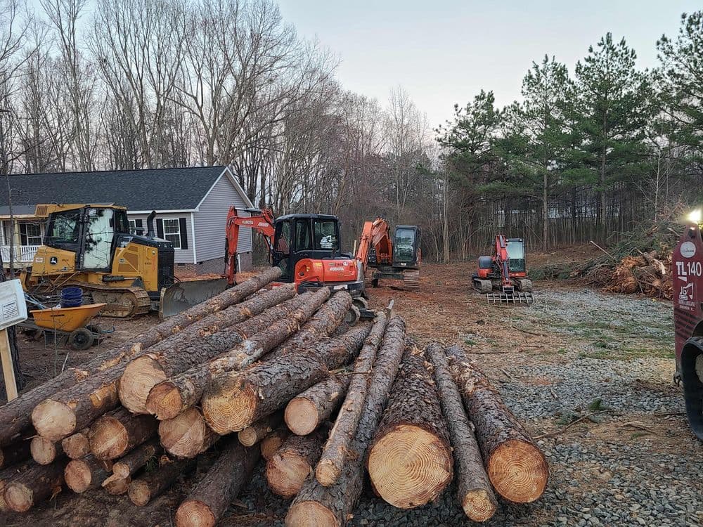Heavy machinery and logs at a construction site surrounded by trees and a house.