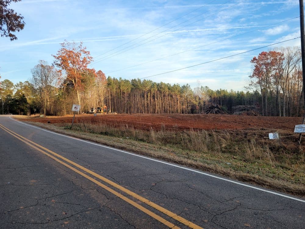 Cleared land by the road with construction equipment and autumn trees in the background.