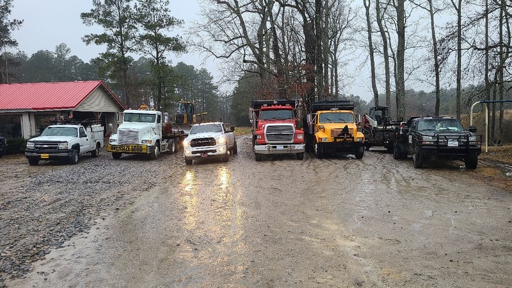Work trucks parked in a muddy yard amidst trees and a building, on a rainy day.