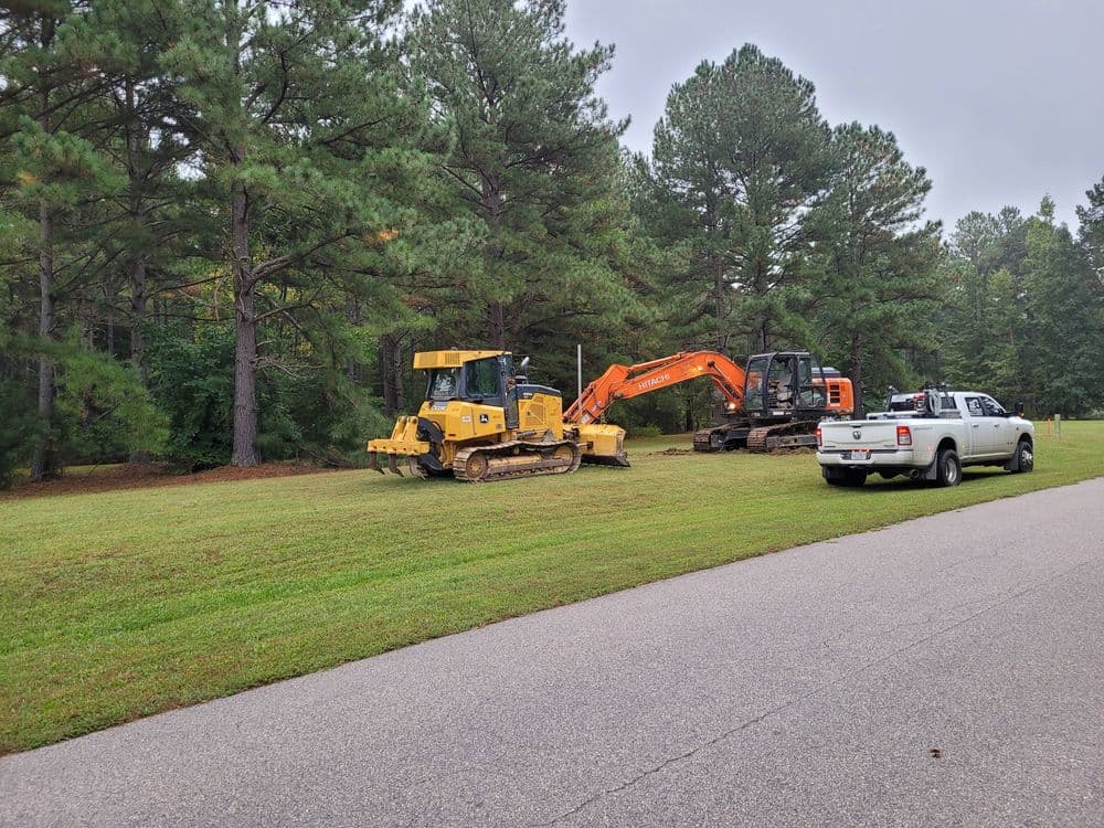 Bulldozer and excavator on a grassy site near a wooded area and parked truck.