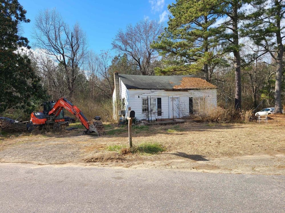 Abandoned house with overgrown yard and construction equipment nearby, rural setting.