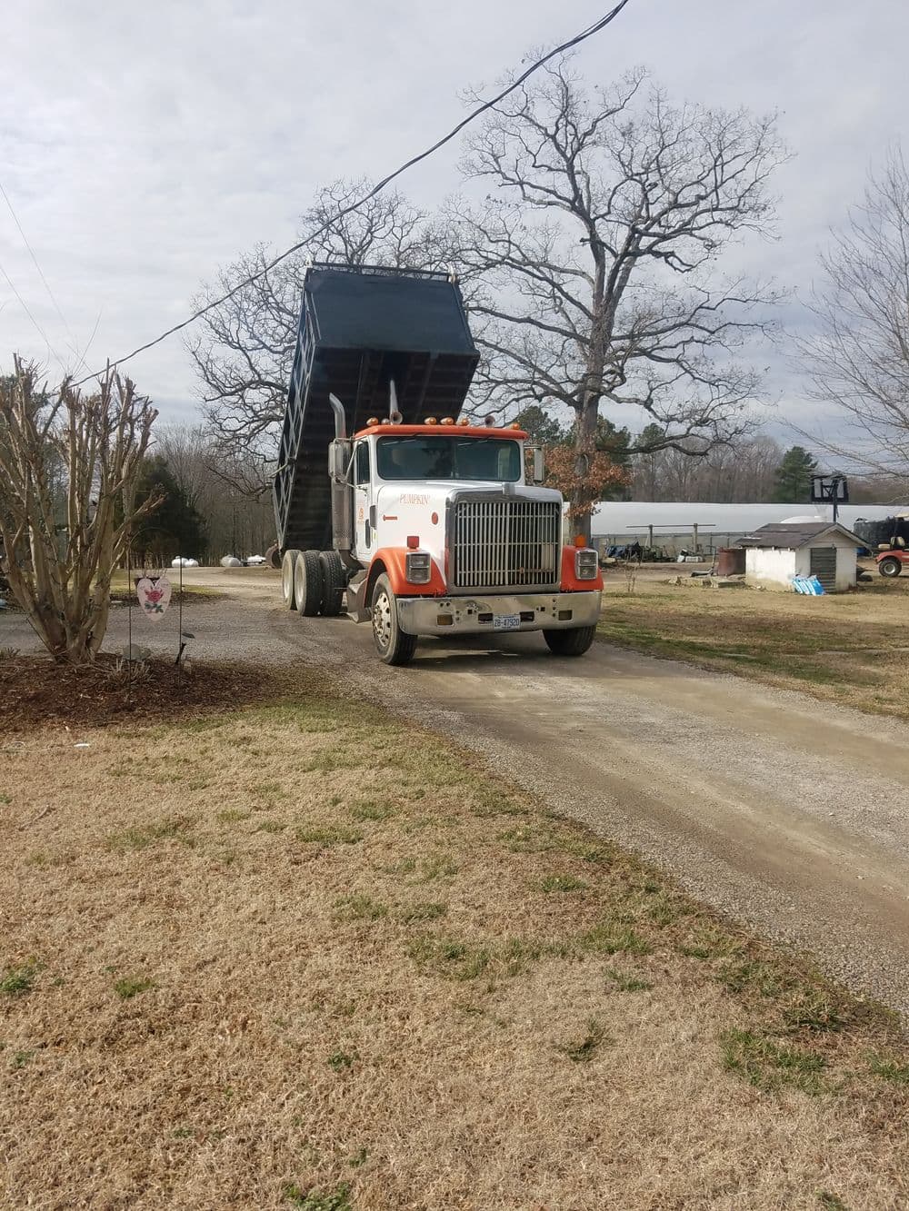 Dump truck unloading materials on a gravel driveway in a rural setting.