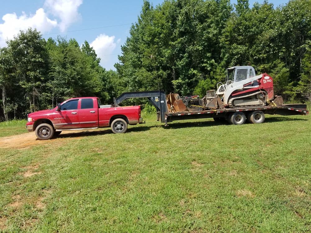 Red pickup truck towing a flatbed trailer with a compact skid steer loader in a grassy field.