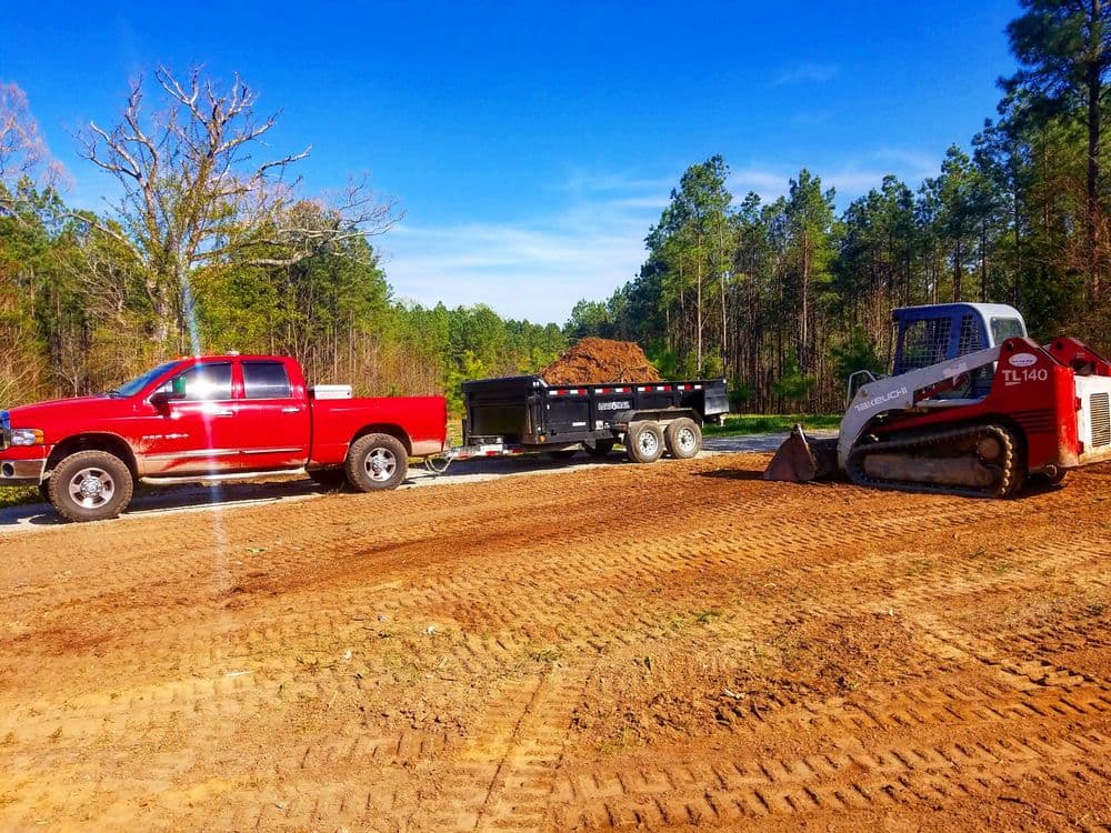 Red truck and trailer with dirt beside a skid steer loader on a construction site.
