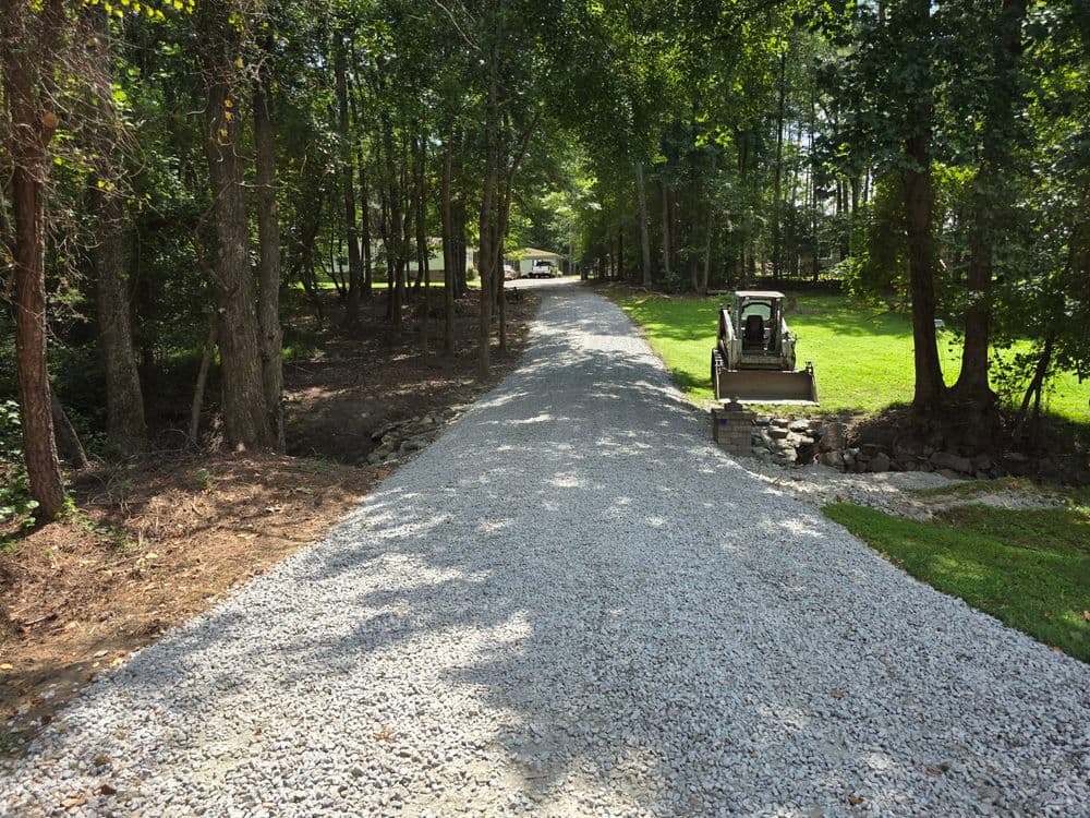 Gravel driveway through wooded area, leading to a house and landscaping.