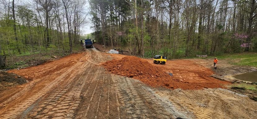 Construction site showing a dirt road being developed, heavy machinery, and workers in a forested area.