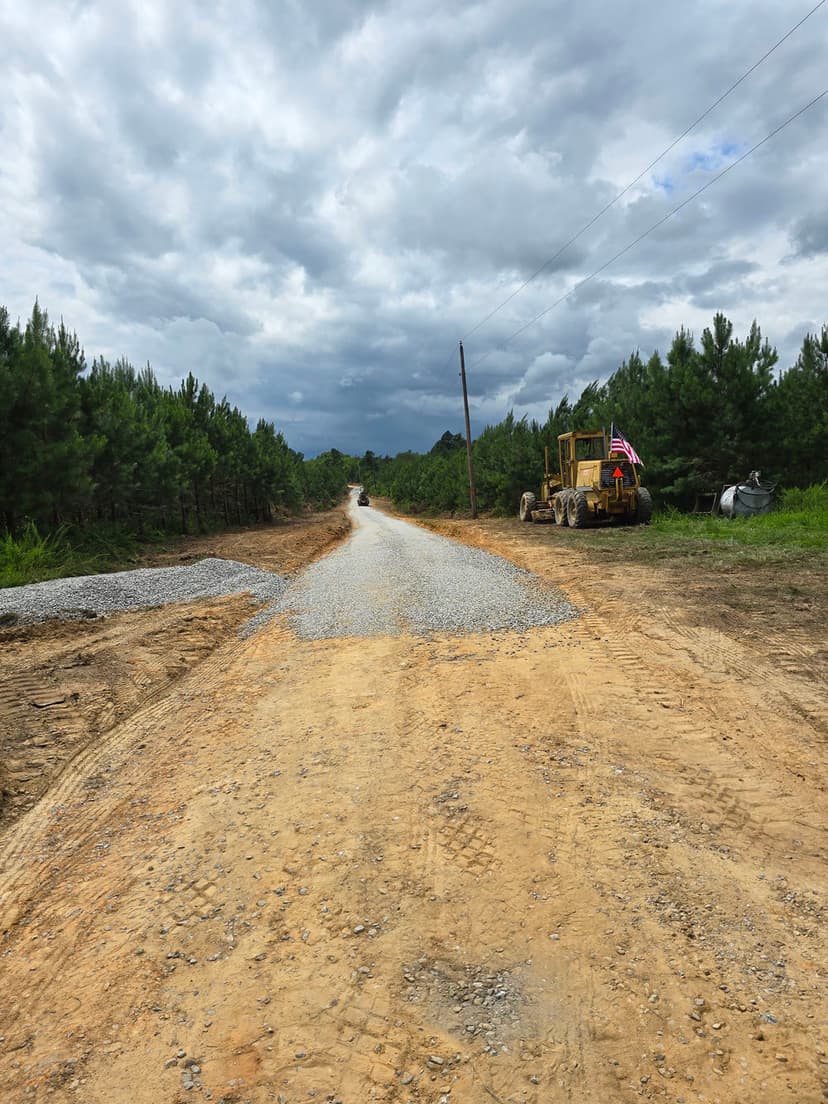 Gravel road construction amidst pine trees under a cloudy sky, construction equipment nearby.