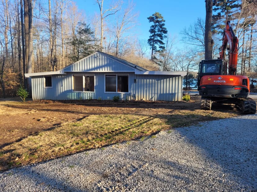 Modern house renovation with excavator, surrounded by trees and freshly landscaped yard.