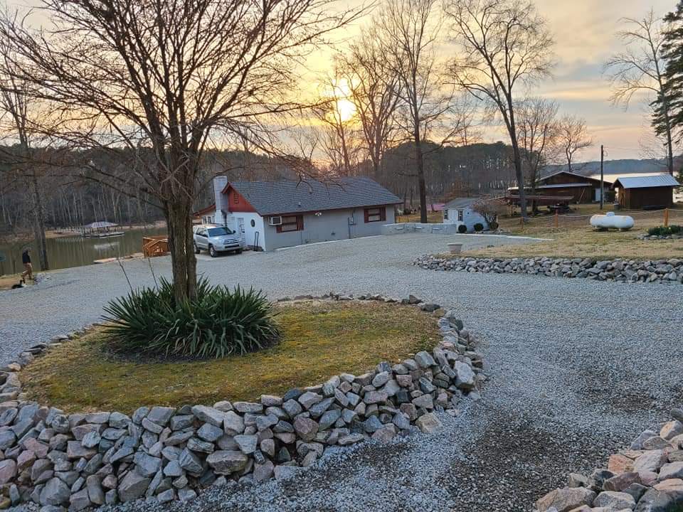 Scenic lake view with gravel driveway, trees, and rustic buildings at sunset.
