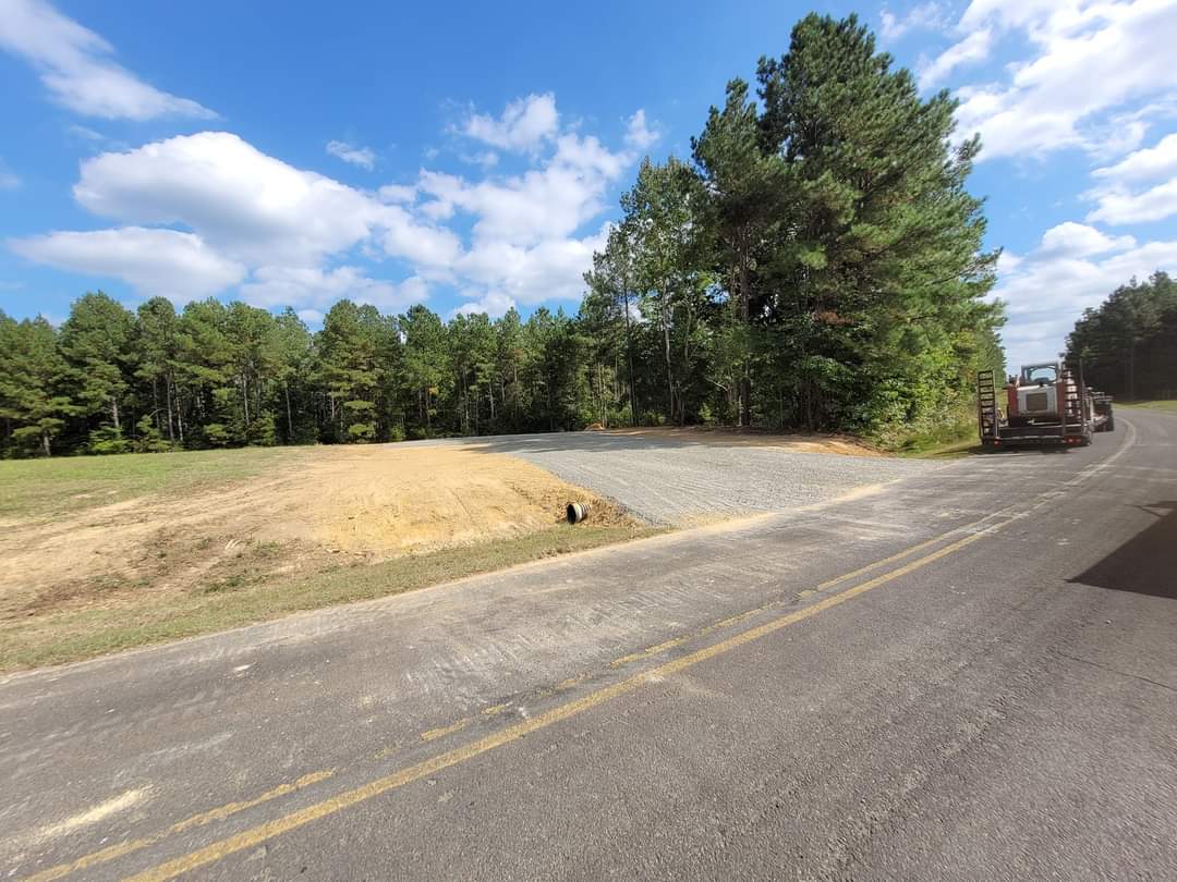Cleared land next to a road, featuring gravel surface and surrounding trees under blue sky.
