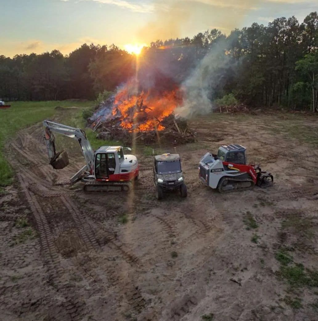 Excavators and equipment near a burning pile of brush at sunset in a clearing.