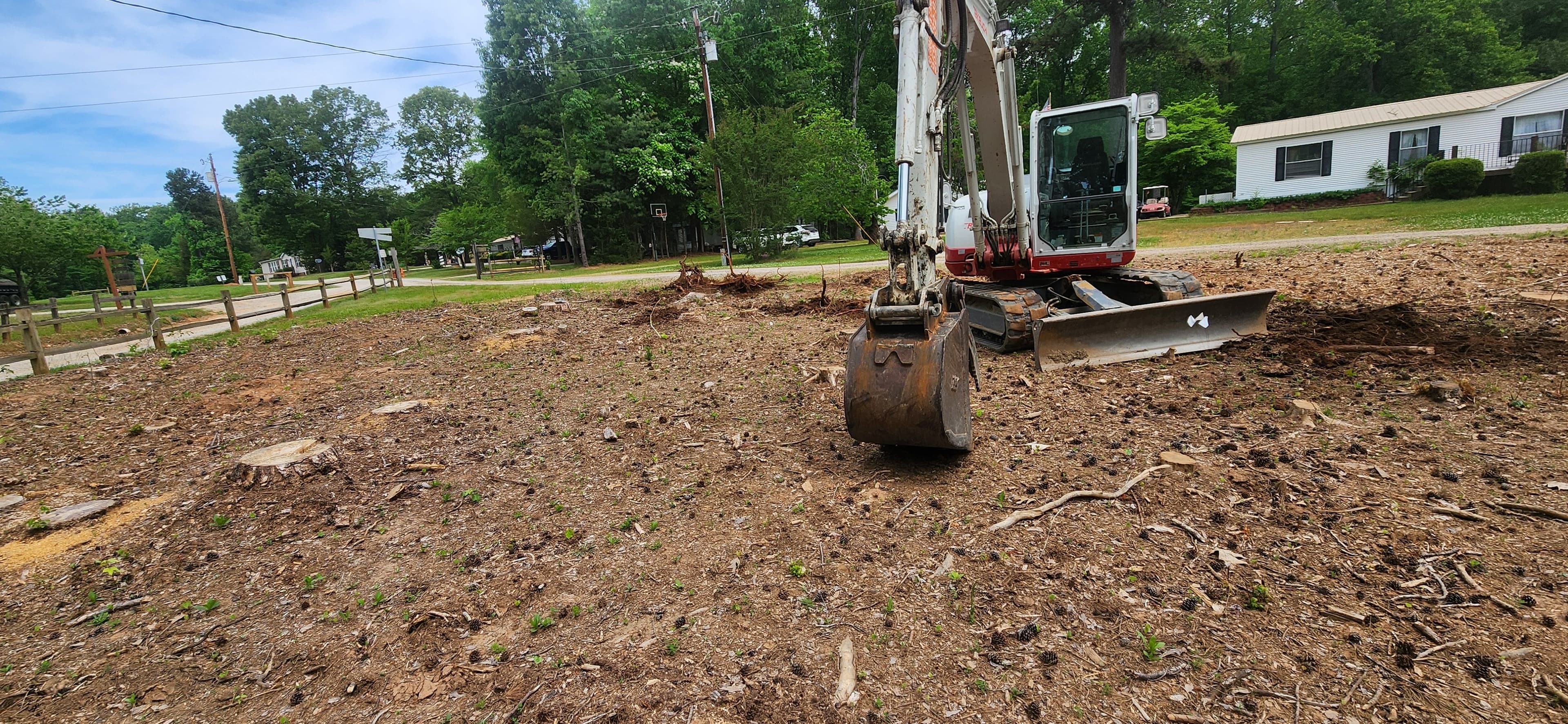 Excavator clearing land with tree stumps in a residential area on a sunny day.