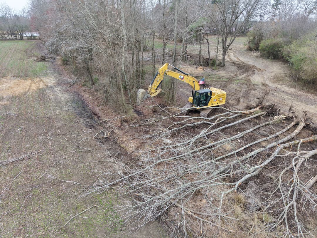 Excavator clearing fallen trees in a rural landscape during land excavation project.