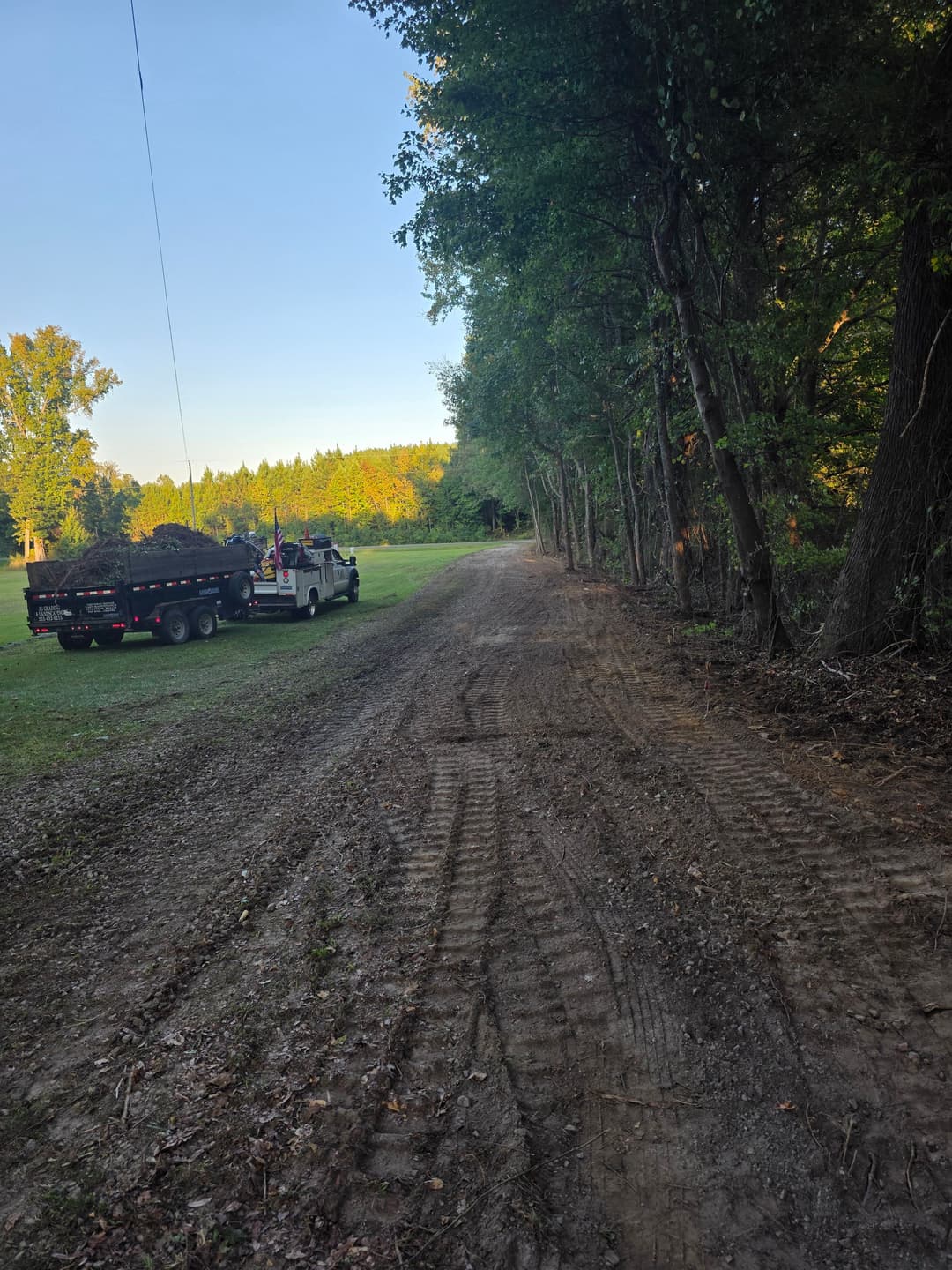 Gravel road lined with trees, truck with trailer parked on the side in a serene landscape.