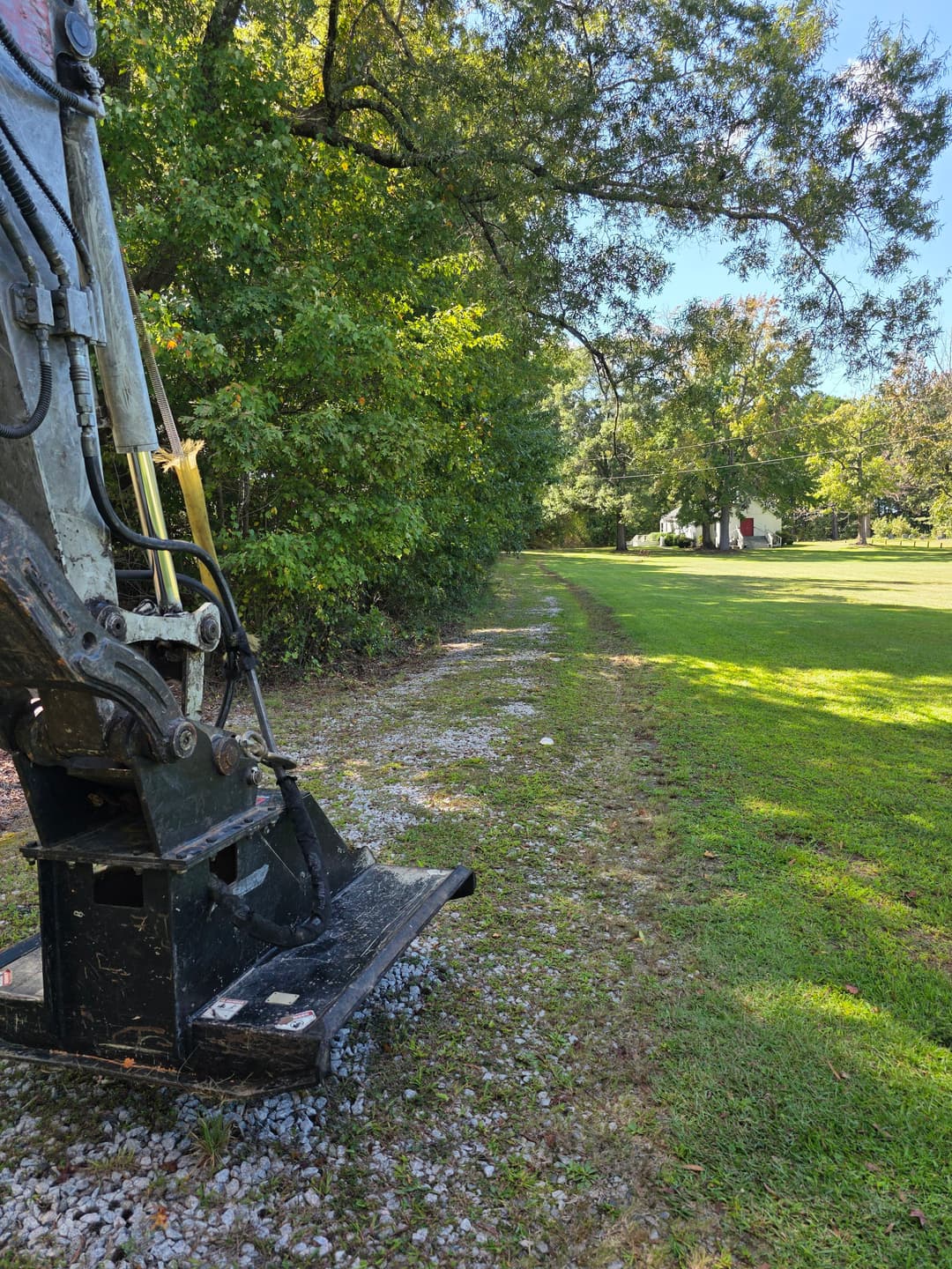 Excavator on gravel path beside trees and open field, sunny day, clear blue sky.
