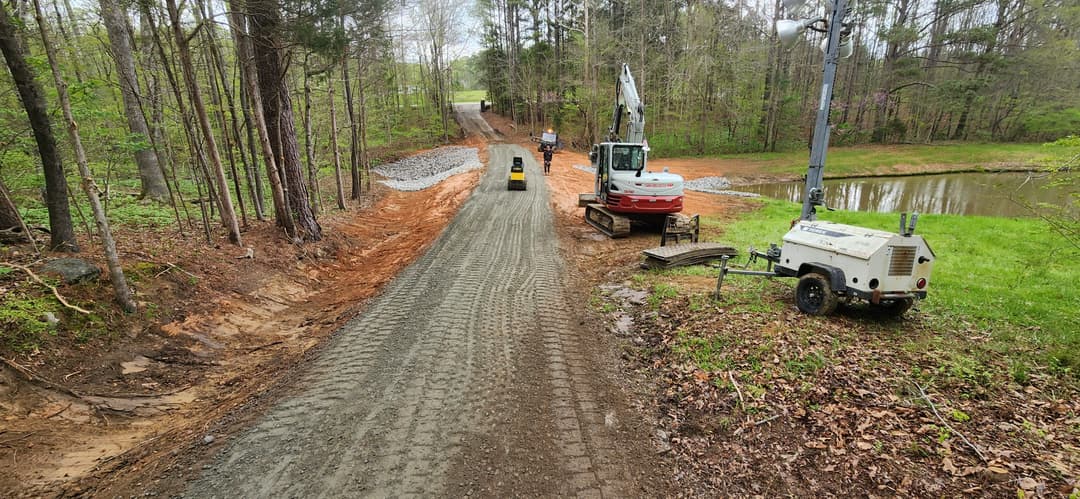 Construction site with machinery and gravel path beside a wooded area and pond.