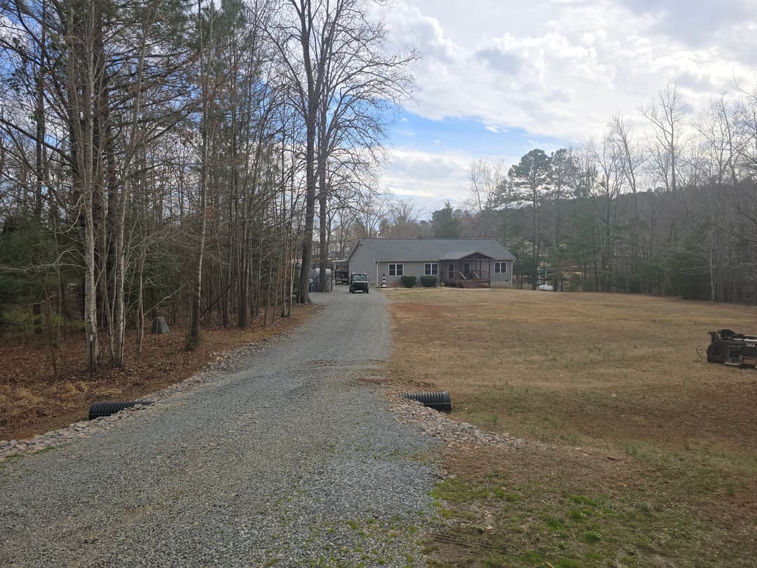 Gravel driveway leading to a house in a wooded area with clear skies and open yard.