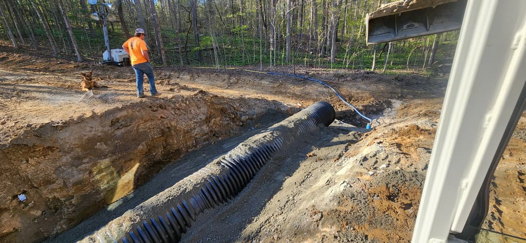 Worker in orange shirt oversees a trench with drainage pipe installation in a wooded area.