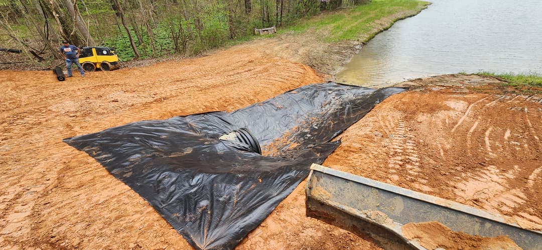 Black plastic cover laid over sandy soil near a pond, with an equipment vehicle nearby.