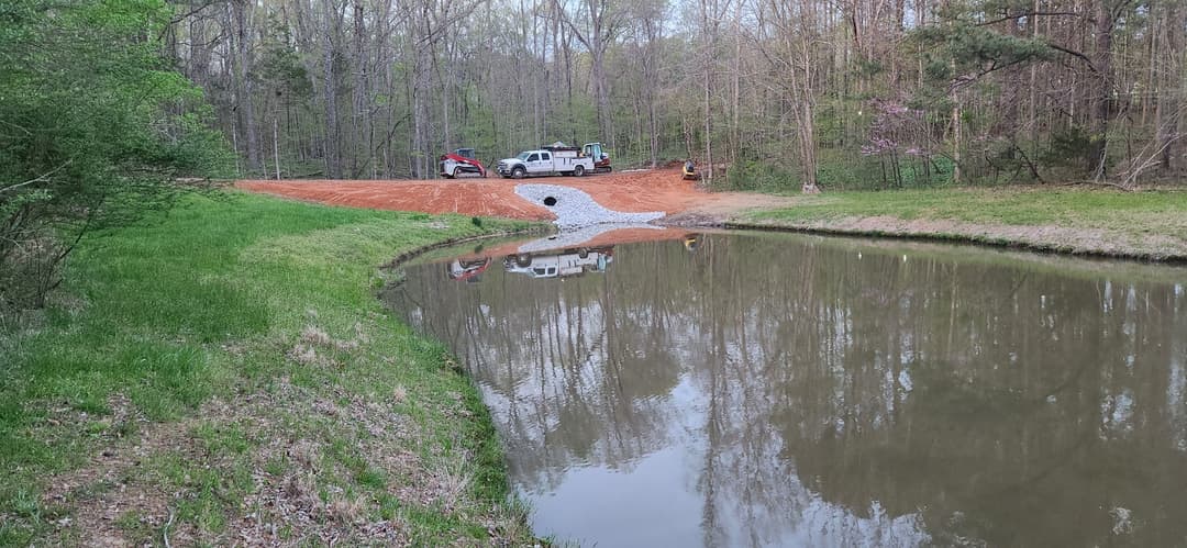 Construction site near a peaceful pond with vehicles in a wooded area.