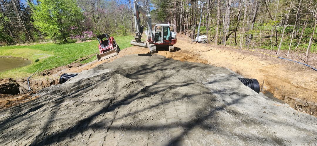 Excavator and machinery working on a dirt path near a wooded area and pond.
