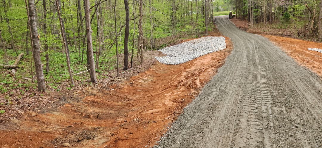 Gravel road construction in wooded area with freshly graded earth and drainage stones.