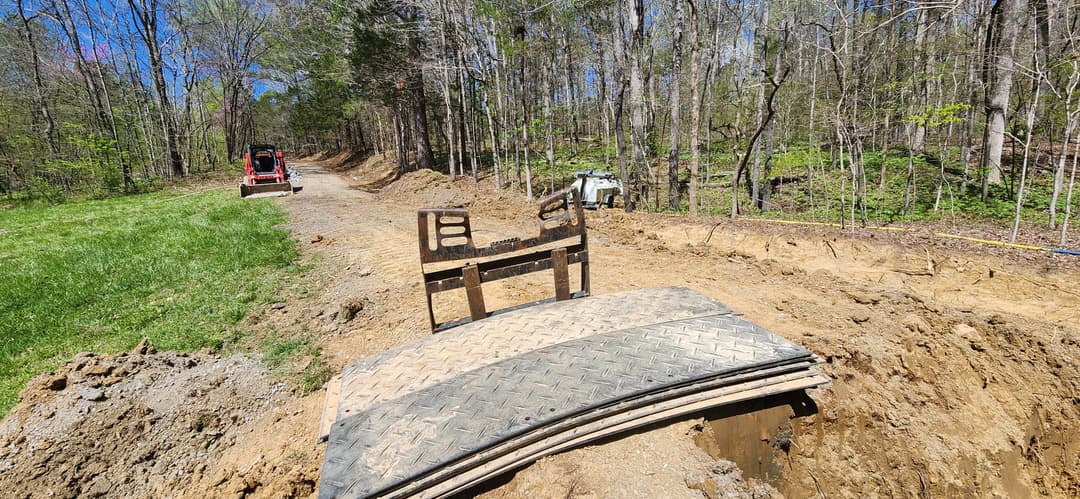 Wooden ramp on dirt path in forested area with construction machinery in the background.